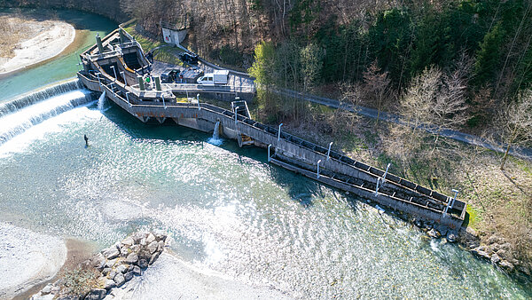 Luftaufnahme eines Wasserkraftwerks und Staudamms an einem Fluss mit überfließendem Wasser. Die Anlage zeigt Plattformen, technische Einrichtungen und Absturzsicherungen zur Wartung.